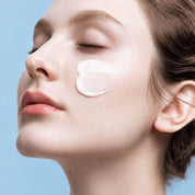 Close-up of a woman with her eyes closed, showing a ANUA Barrier Reboot Intensive Cream applied to her cheek against a light blue background.