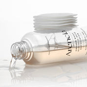 A clear glass bottle of  ARENCIA Eraser Shot Glycolic Acid Exfoliator Emulsion with pale peach liquid, shown lying on its side with liquid dripping out, alongside a stack of white cotton pads on a reflective white surface.