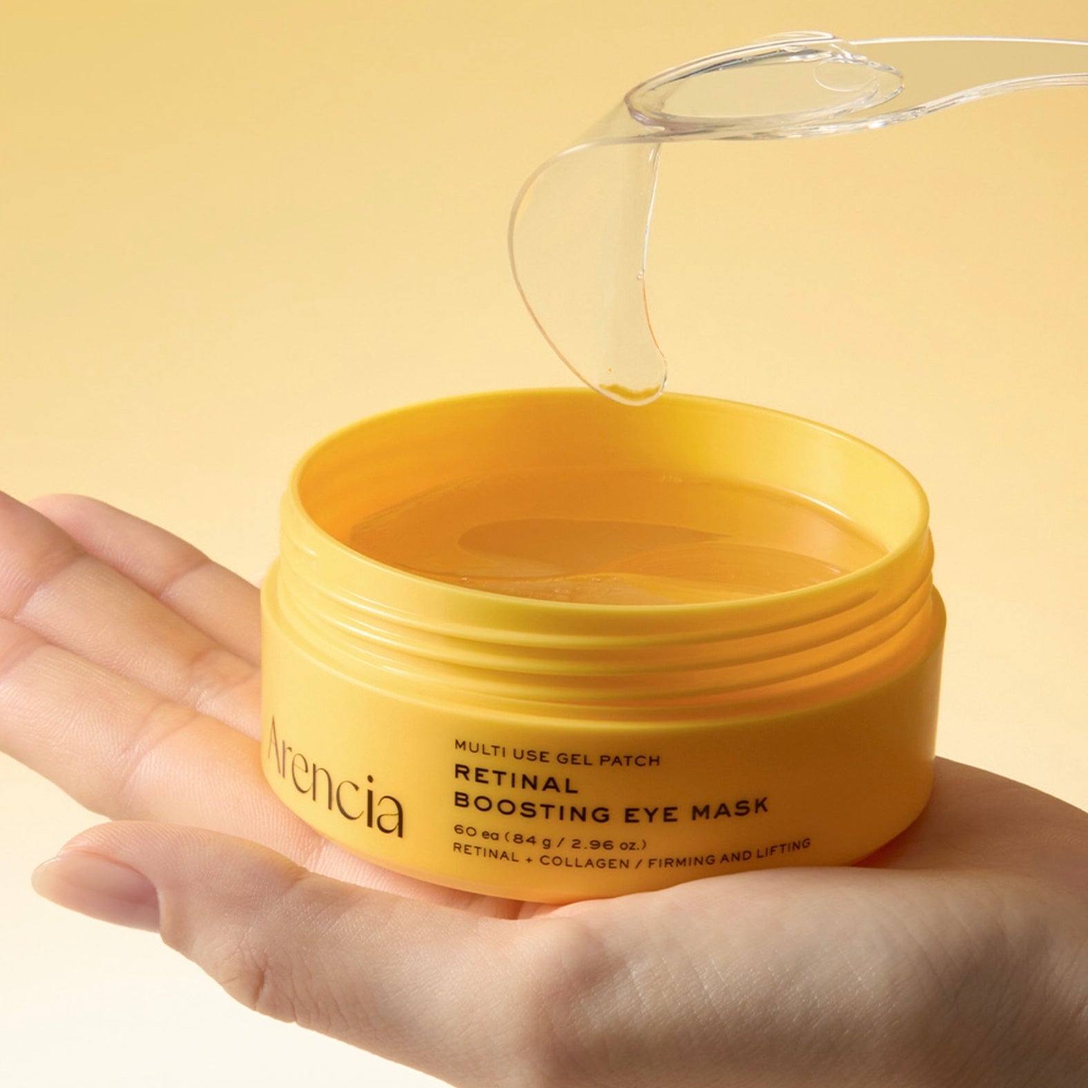 A hand holding an open yellow jar of ARENCIA Retinal Boosting Eye Mask, with a clear gel patch being lifted above the container against a soft yellow background.