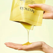 A hand holds a yellow ARENCIA Vita Collagen Radiance Jello Mask to pour a thick, glossy golden-yellow gel into another hand, against a pale yellow background.