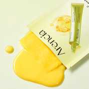 ARENCIA Vitamin C Booster Shot Mask, a tube of serum, and a dollop of bright yellow treatment serum on a clean off-white background.