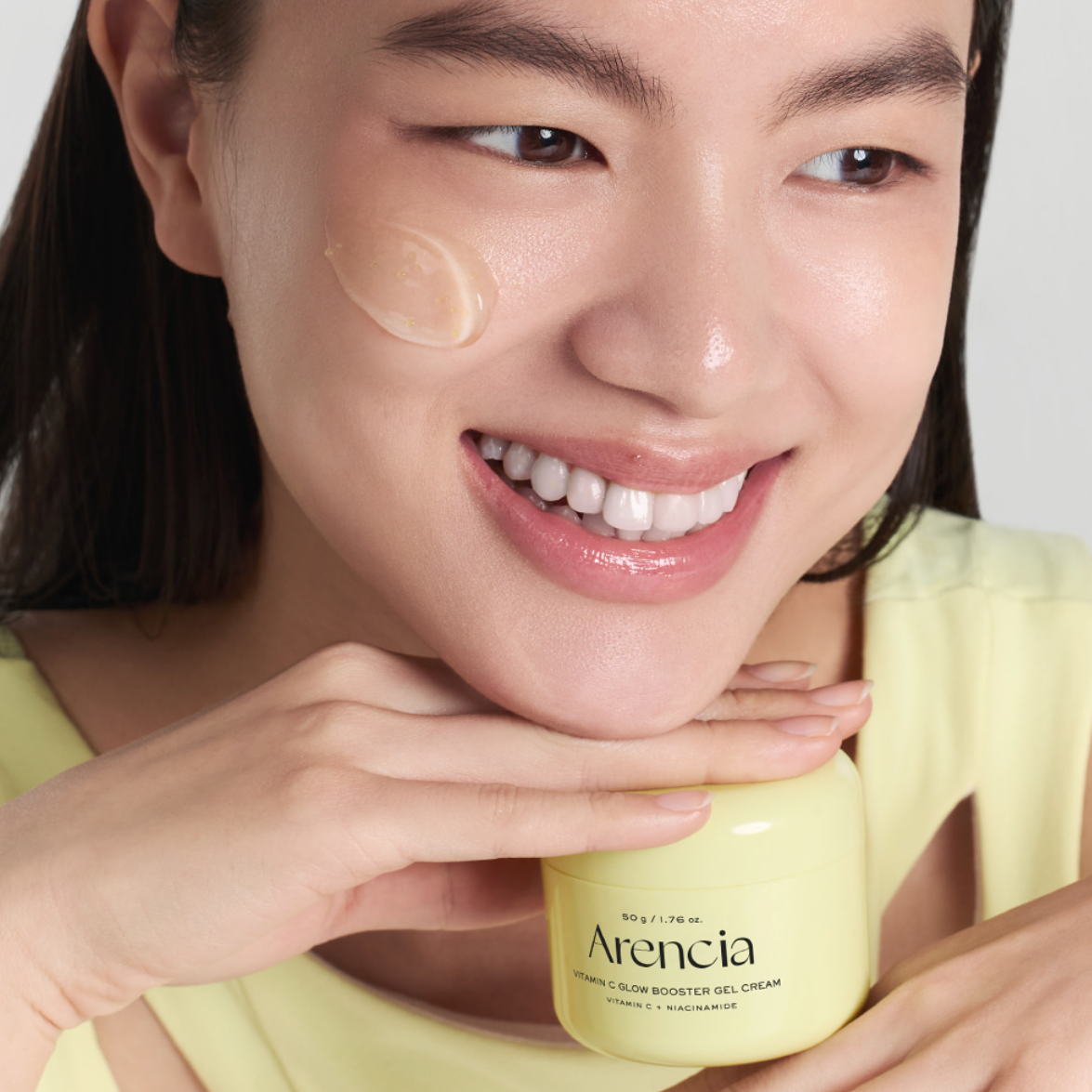A close-up of a smiling woman holding an Arencia Vitamin B3 & Glow Booster Gel Cream jar, with a dollop of the cream visible on her cheek against a clean light gray background.