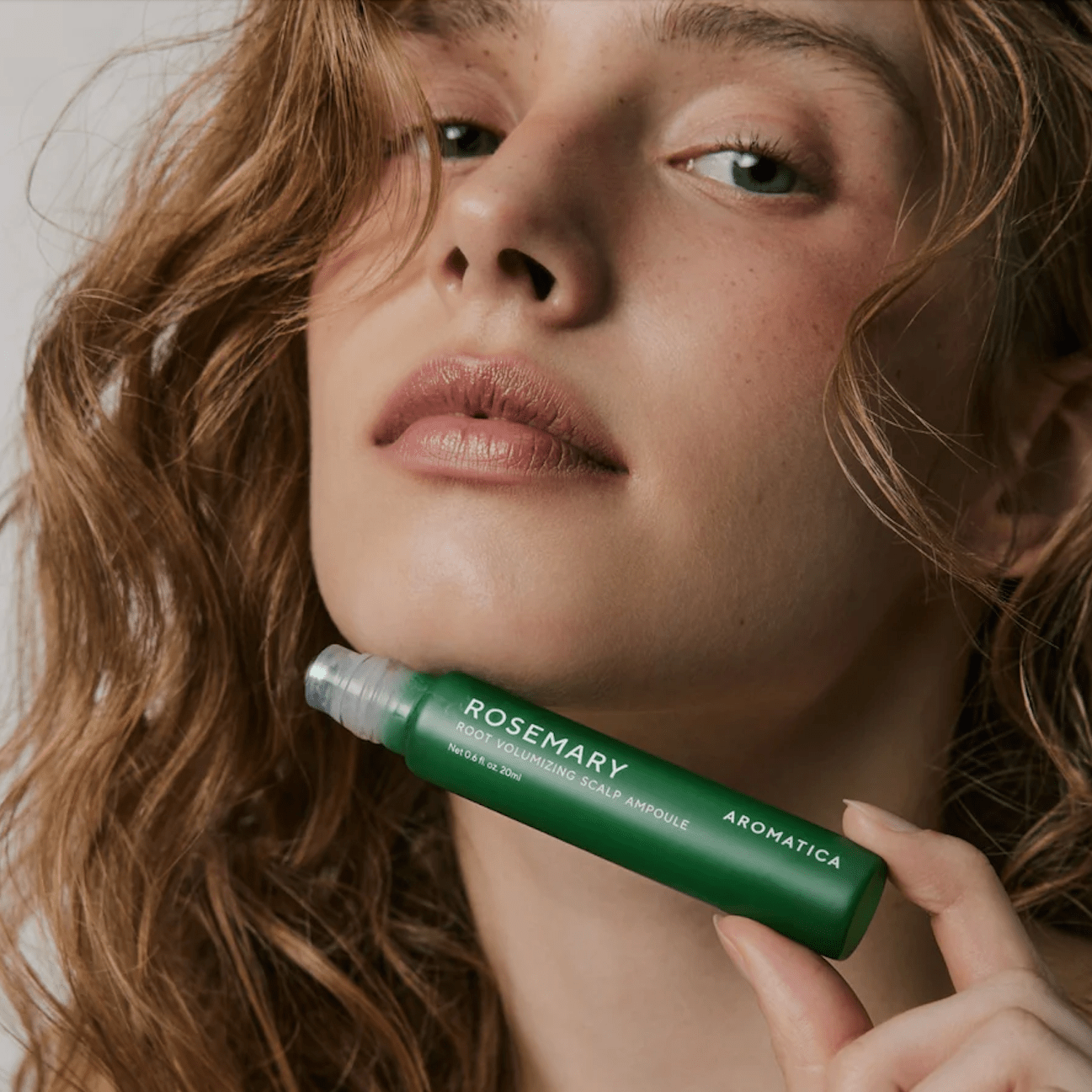 Woman holding a AROMATICA Rosemary Root Volumizing Scalp Ampoule rollerball with 'Rosemary' labeled, against a neutral background.