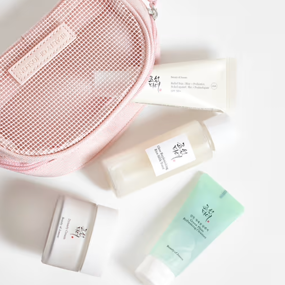 A flat-lay arrangement of Beauty of Joseon Glow Skin Essential Kit (4-Piece Gift Set). on a white background, featuring a pink mesh cosmetic pouch and four white and mint green bottles and jars from the brand Botulose.