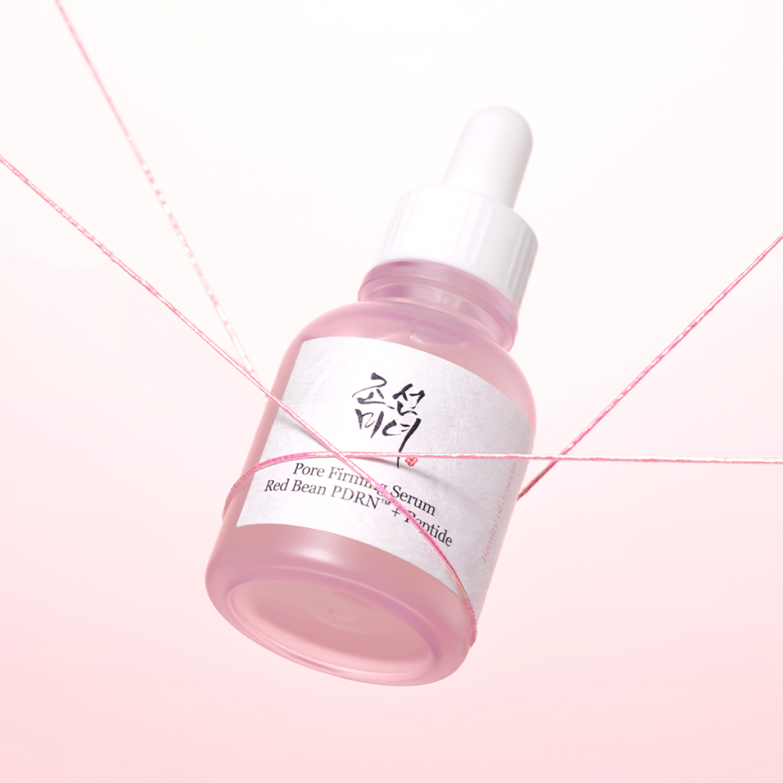 A small dropper bottle of BEAUTY OF JOSEON Pore Firming Serum Red Bean PDRN+Peptide and white cap, shown on a soft pink background with decorative pink threads crossing the frame.