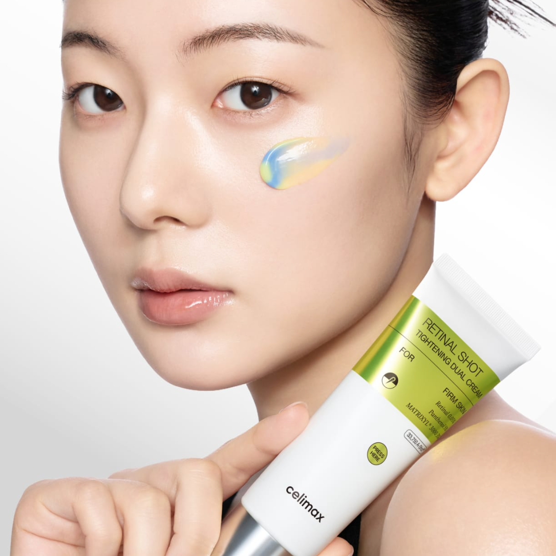 Close-up of a woman holding a tube of CELIMAX The VITA-A Retinal Shot Tightening Dual Cream (50 ml), with a smear of the blue and yellow cream visible on her cheek against a clean white background.