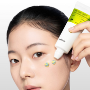 Close-up of a woman applying CELIMAX The VITA-A Retinal Shot Tightening Dual Cream (50 ml), with a hand holding the white and yellow tube against her temple and two small dollops of cream on her cheek.
