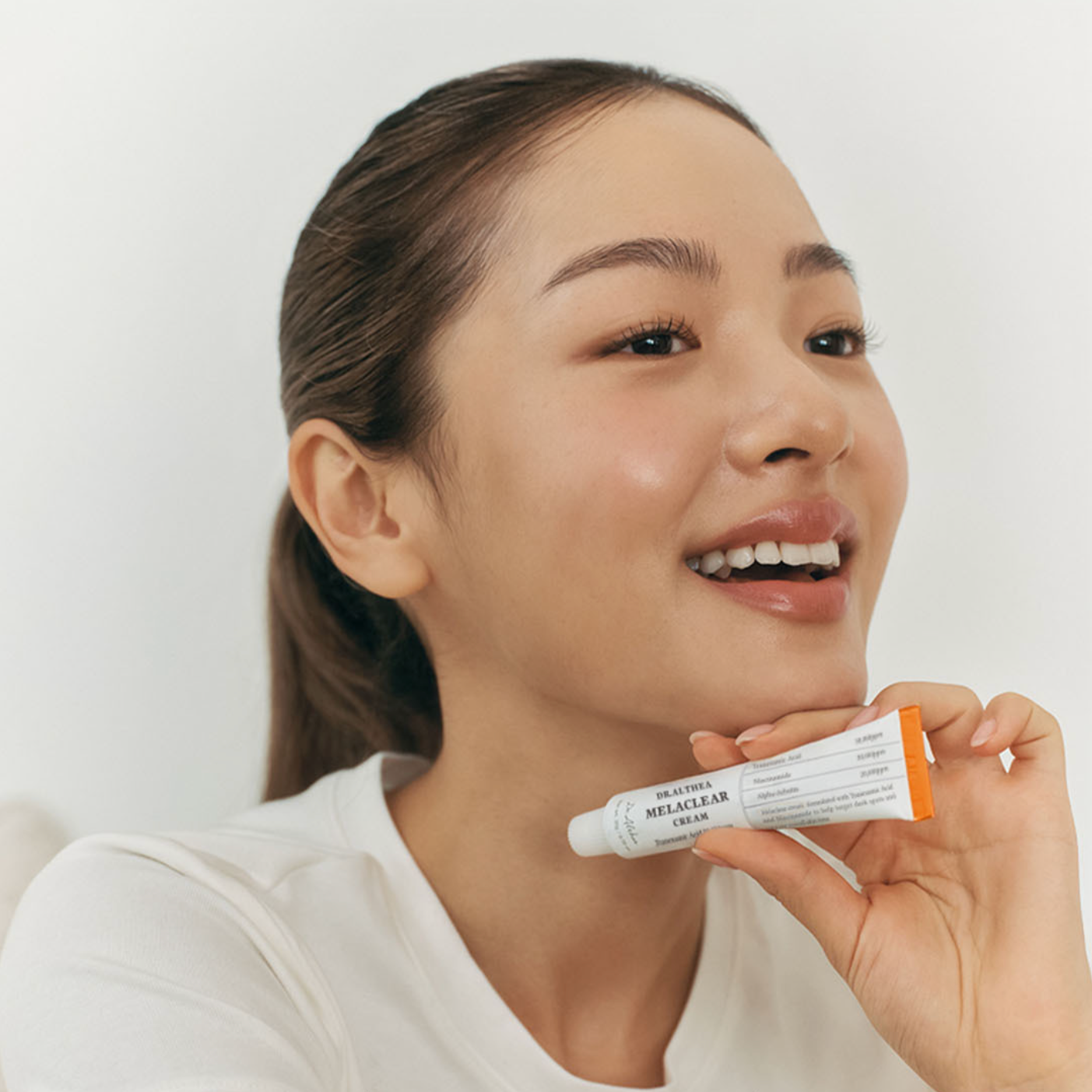 A woman with her hair pulled back holds a white tube of Dr althea makeup against a clean white background.