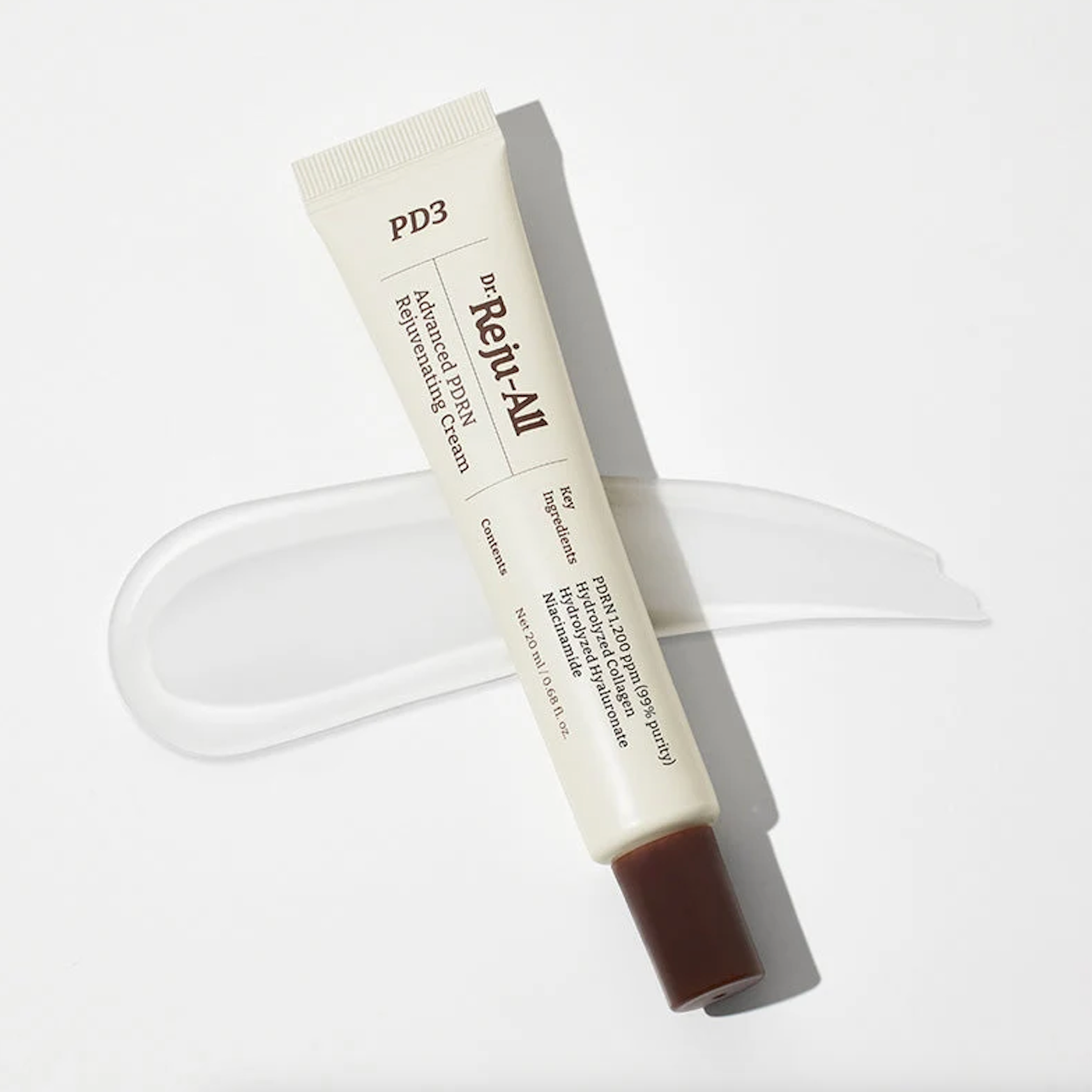 A tube of Dr. Reju-All Advanced PDRN Rejuvenating Cream lies diagonally on a clean white background, placed over a translucent smear of the white cream product. The tube is off-white with black text and a dark brown cap.