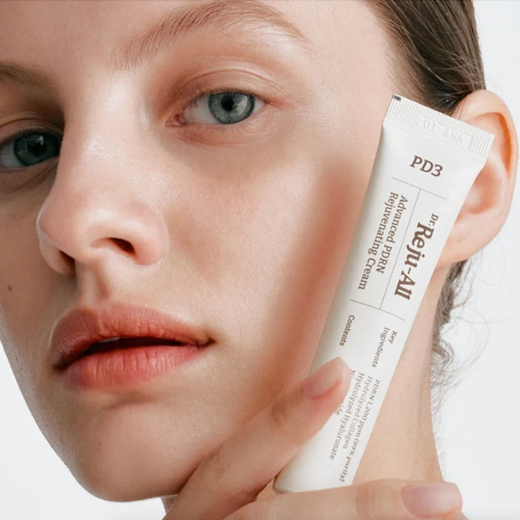 Close-up of a woman holding a white tube of Dr. Reju-All Advanced PDRN Rejuvenating Cream against her cheek, showing the product packaging and texture.