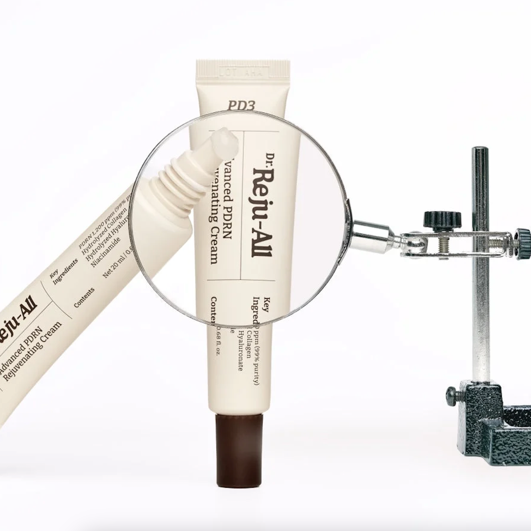 A cream-colored tube of Dr. Reju-All Advanced PDRN Rejuvenating Cream with a dark brown cap, shown with a magnifying glass graphic and a second angled view of the tube on a white background.