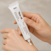 A close-up of a person's hands holding a white tube of Dr. Reju-All Advanced PDRN Rejuvenating Cream, with a small dollop of cream squeezed out at the tip, against a soft beige background.