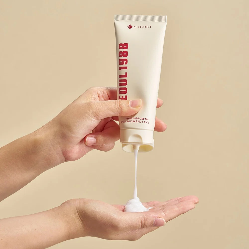 Hand holding a tube of K-SECRET Seoul 1988 Cream: Snail Mucin 93% + Rice with visible cream on palm against a beige background