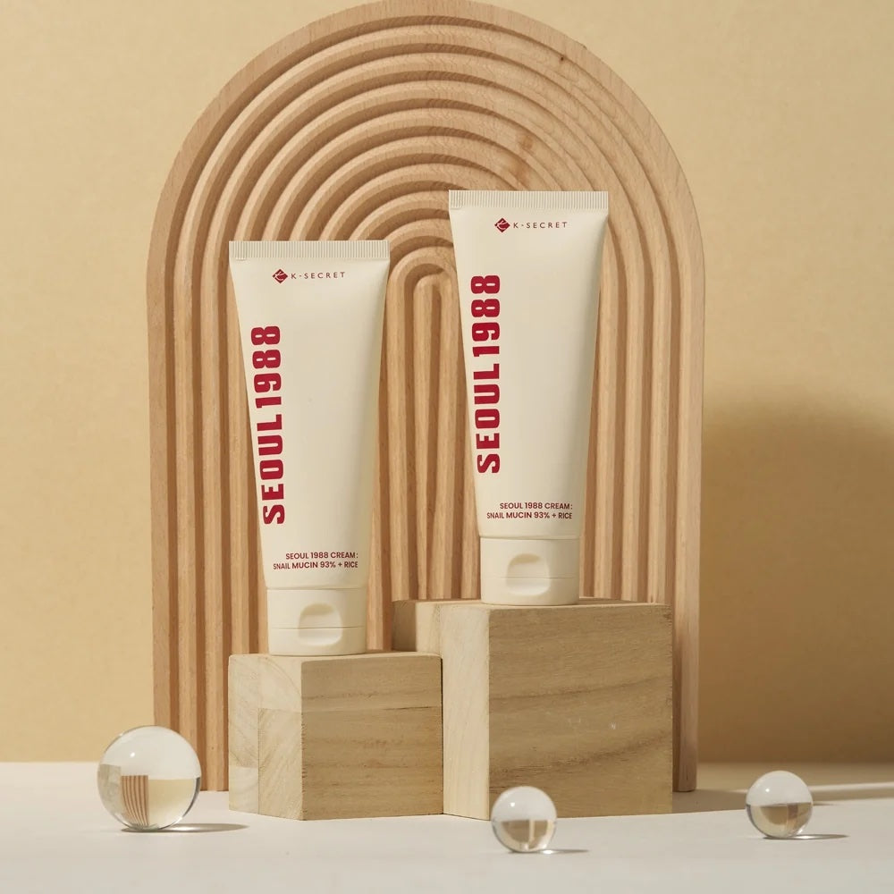 Two white cosmetic tubes labeled K-SECRET Seoul 1988 Cream: Snail Mucin 93% + Rice stand on wooden blocks in front of a beige arched backdrop. The tubes appear to be skincare cream products, photographed in a styled studio setup with small glass spheres on a white surface.