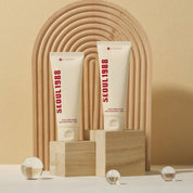 Two white cosmetic tubes labeled K-SECRET Seoul 1988 Cream: Snail Mucin 93% + Rice stand on wooden blocks in front of a beige arched backdrop. The tubes appear to be skincare cream products, photographed in a styled studio setup with small glass spheres on a white surface.