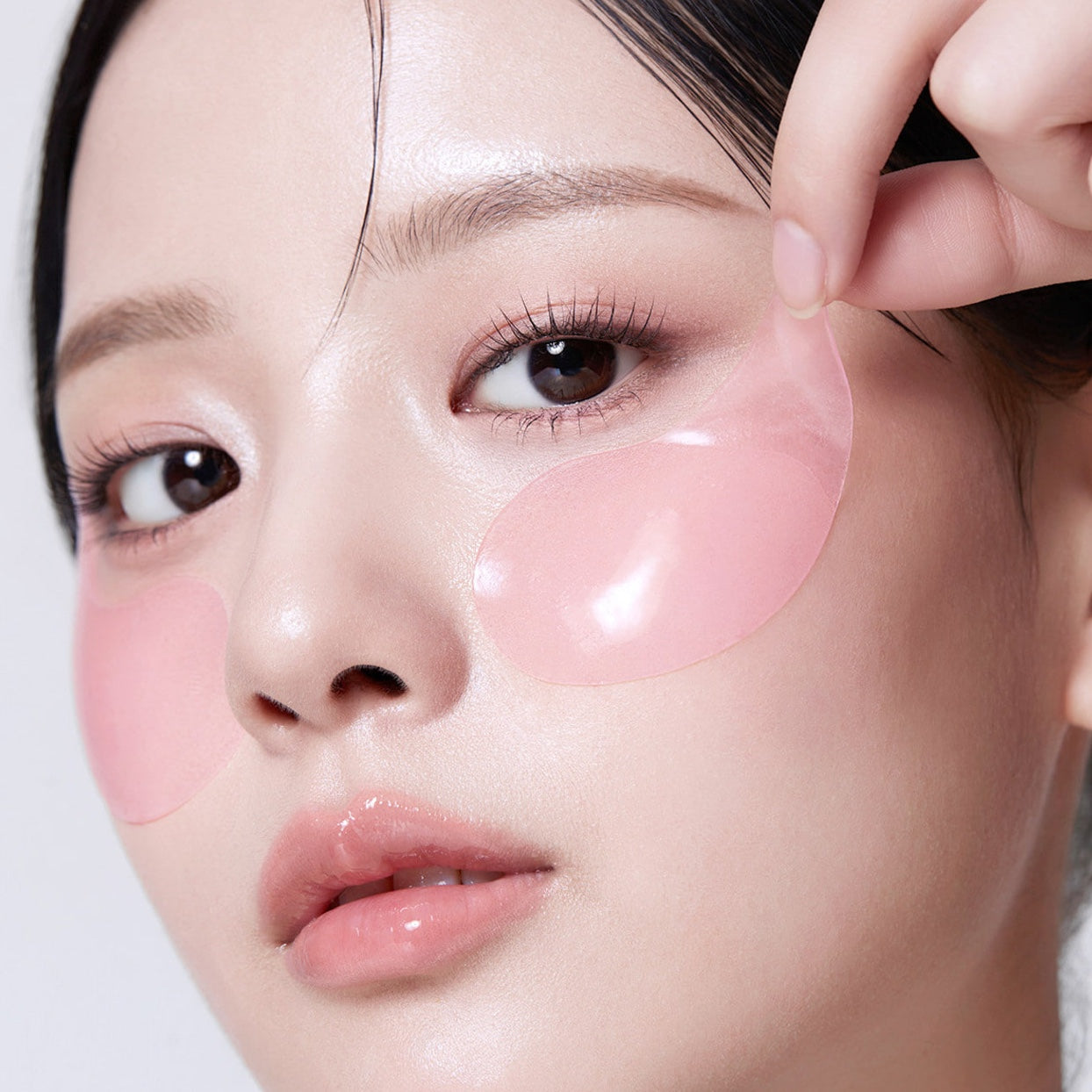 Close-up of a woman applying a translucent pink under-eye patch to her left eye while another pink patch is visible on her right eye.