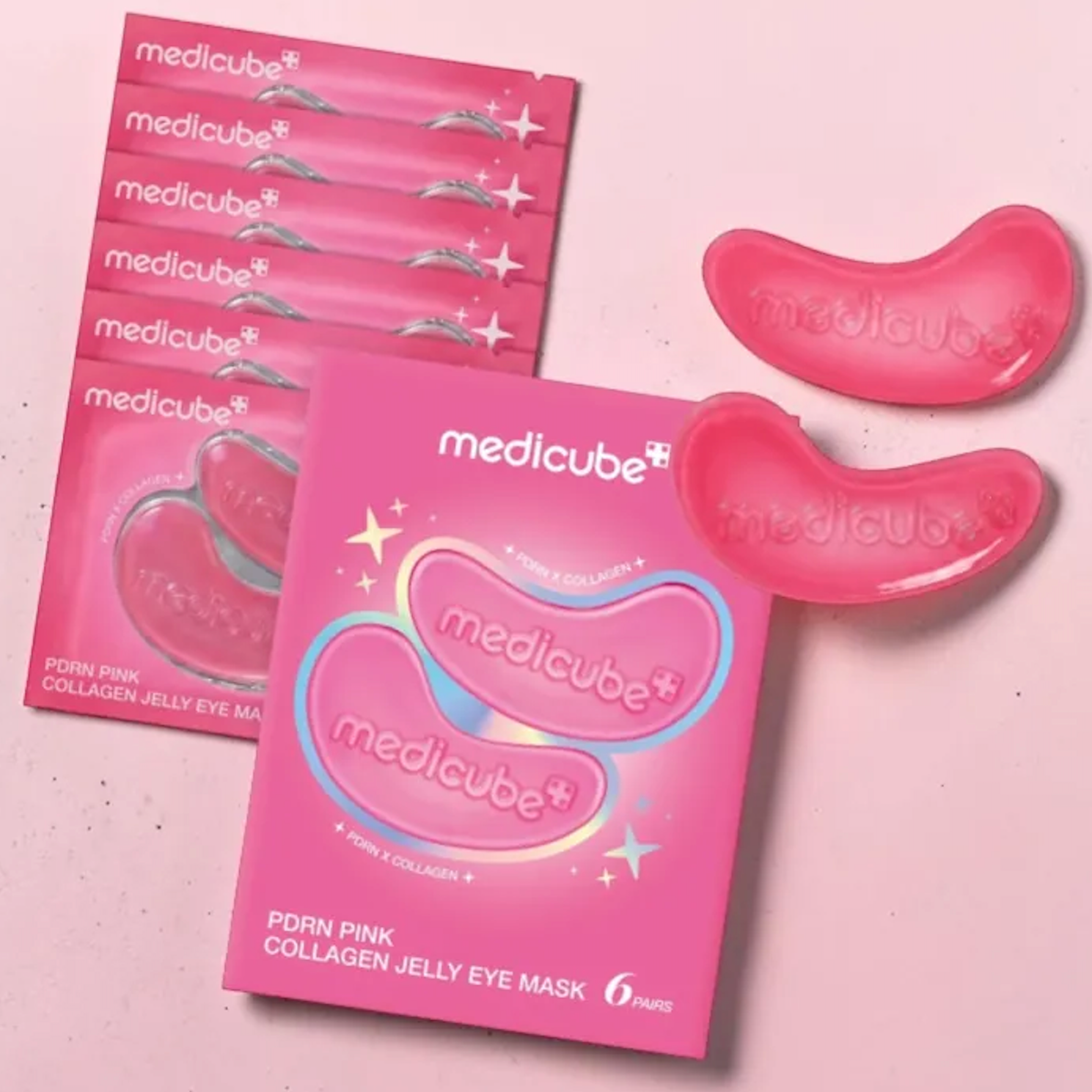 Six pink packets of Medicube PDRN Pink Collagen Jelly Eye Masks are arranged on a light pink background, with two translucent pink jelly eye masks displayed to the right. The packaging features white and blue branding and text.