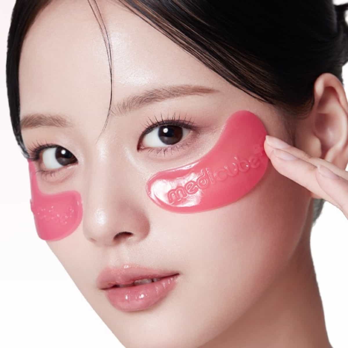 Close-up of a woman wearing MEDICUBE PDRN Pink Collagen Jelly Eye Mask (6 Pouch) under her eyes, with one patch being peeled back by a hand, on a clean white background.