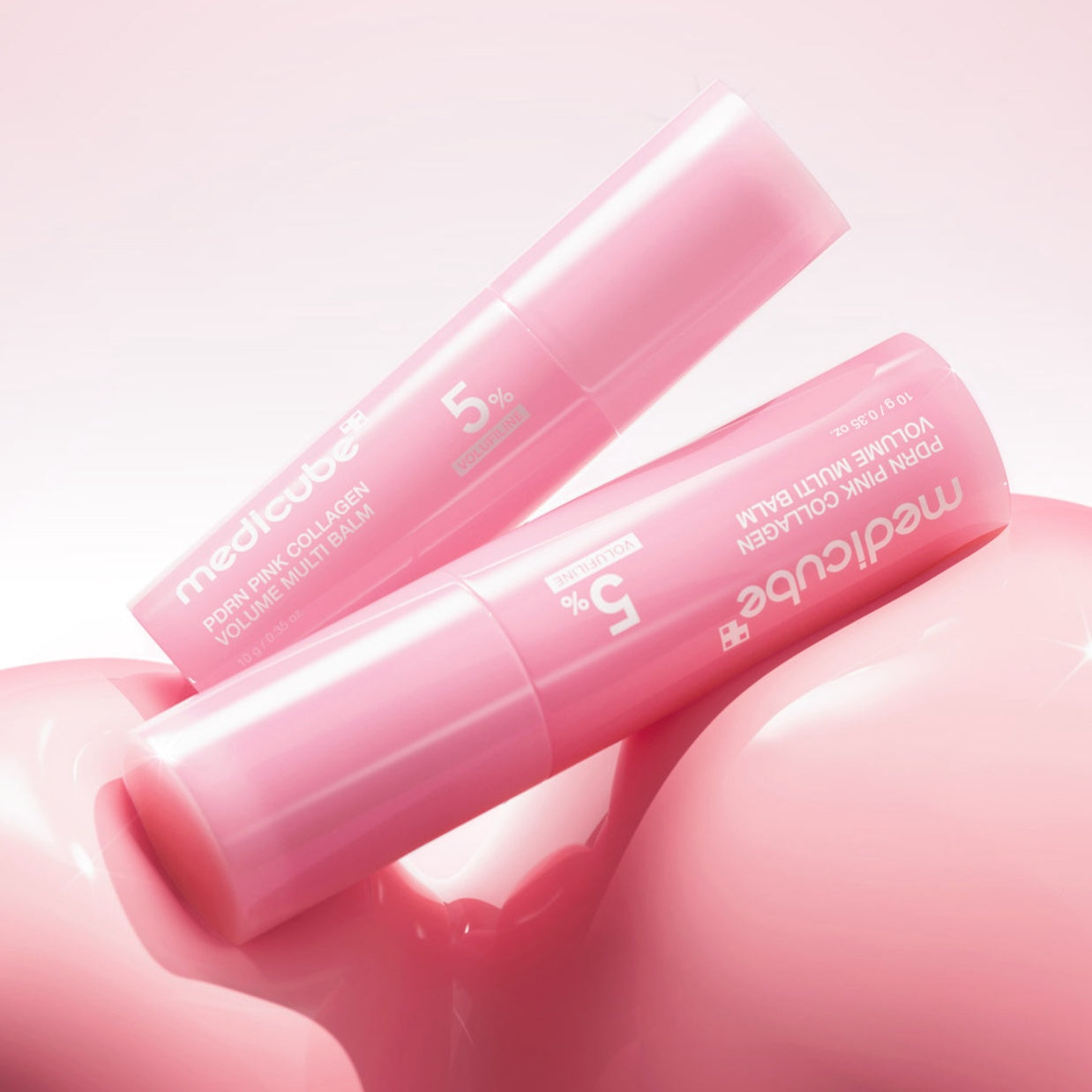 Two pink MEDICUBE PDRN Pink Collagen Volume Multi Balm sticks resting on a glossy pink surface against a soft pink background.