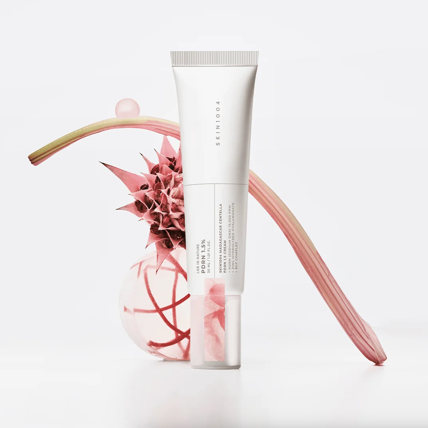 A white cosmetic tube labeled SKIN1004 Madagascar Centella PDRN 1.5 Cream stands upright on a white surface, styled with pink botanical props against a clean white background.