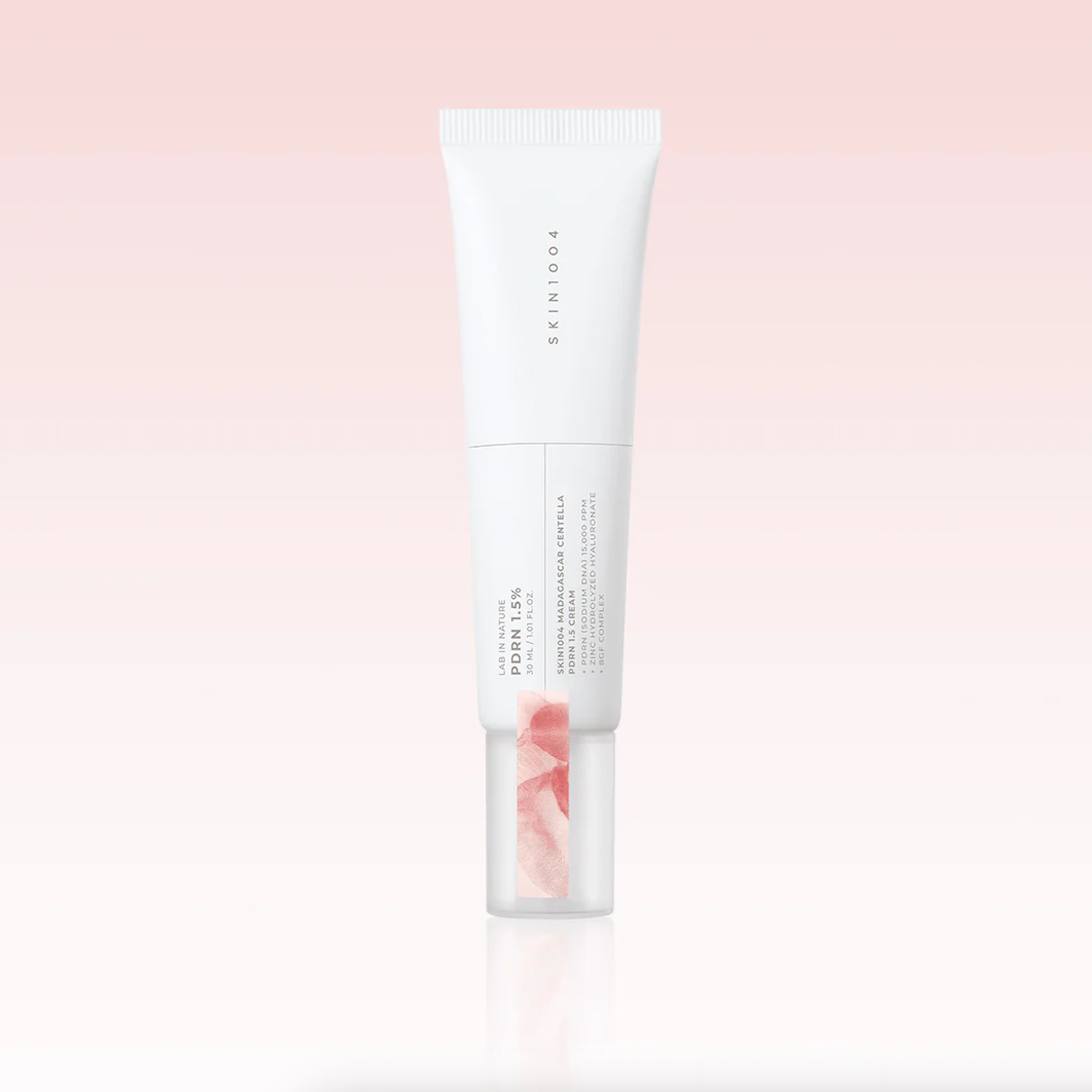 A white squeeze tube of SKIN1004 Madagascar Centella PDRN 1.5 Cream with a clear cap showing pink serum inside, centered on a soft pink gradient background.