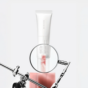 SKIN1004 Madagascar Centella PDRN 1.5 Cream stands upright on a pink block, with a circular magnifying glass overlay centered on the tube. The setup is photographed against a very light gray/white studio background with metallic stand hardware visible at the bottom.