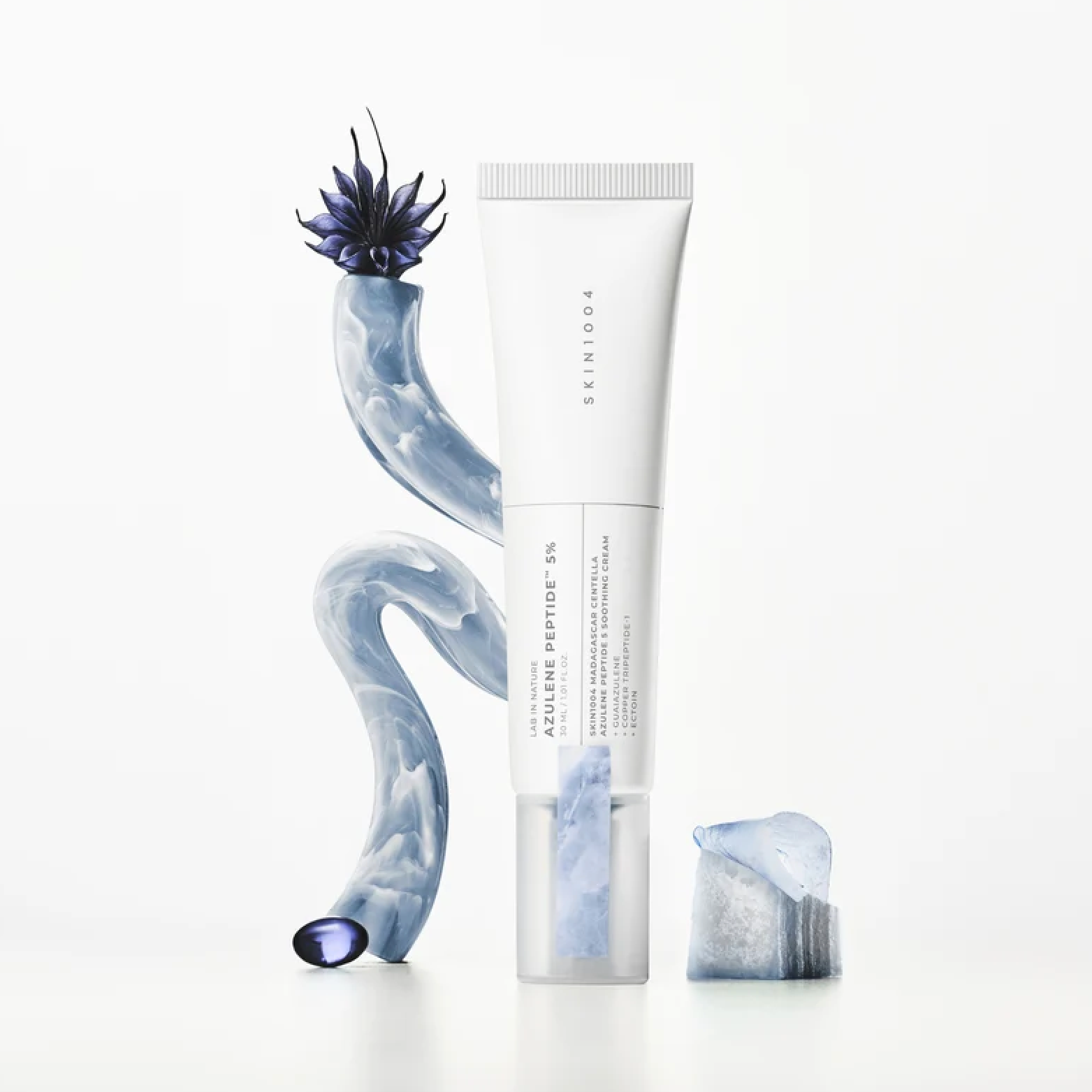 A white skincare tube labeled SKIN1004 Madagascar Centella Azulene Peptide 5 Soothing Cream stands upright on a clean white background, flanked by sculptural blue marbled props and a small clear cap resting beside it.